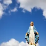 Mali kip Gospe u bijeloj haljini i plavom plaštu s raširenim rukama ispred bijelih oblaka i sunčanog neba. mojmolitvenik.com